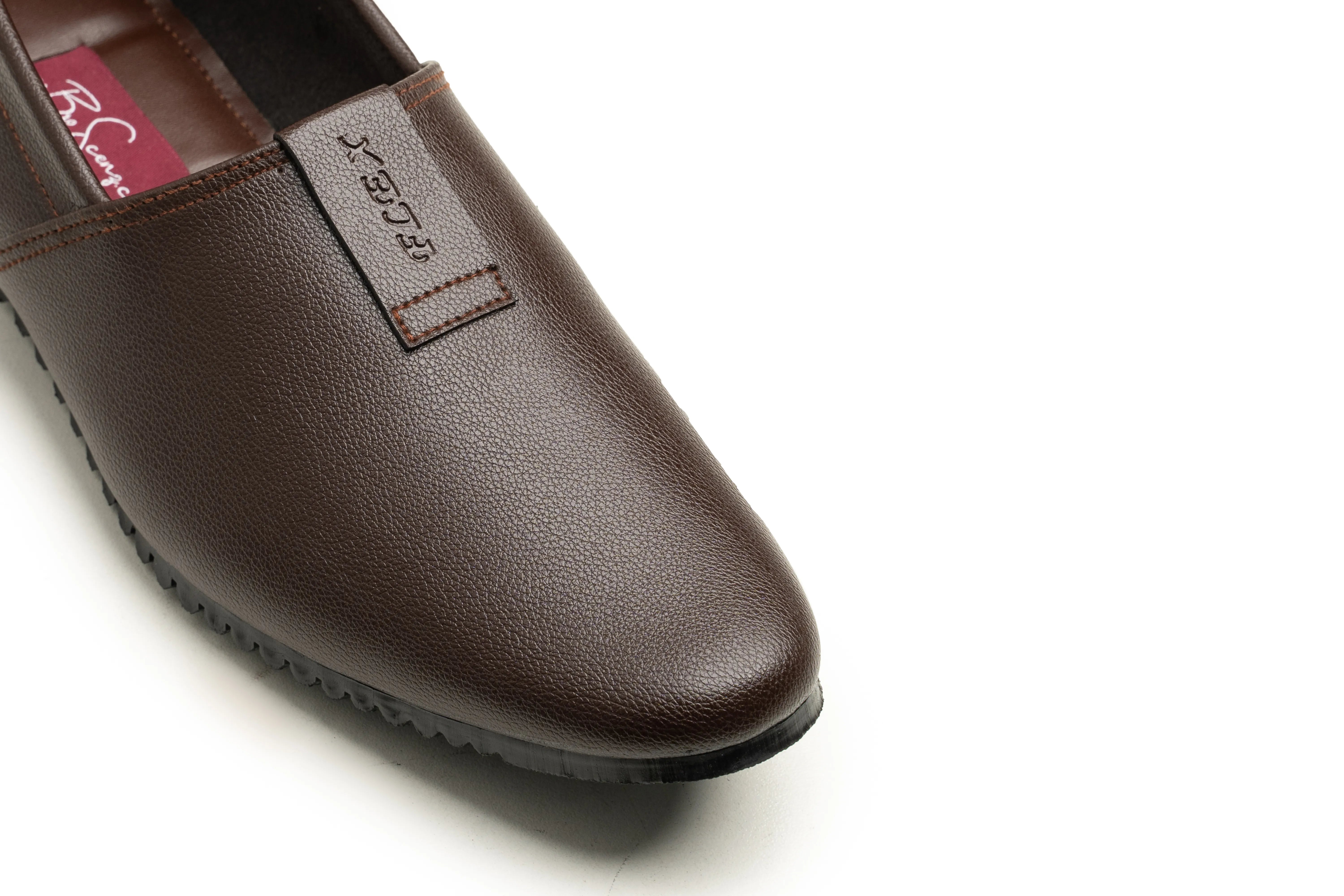 A close-up of a dark brown leather slip-on shoe, showing the textured leather upper, a matching brown pull tab with debossed branding, and a black outsole, photographed on a clean white background.