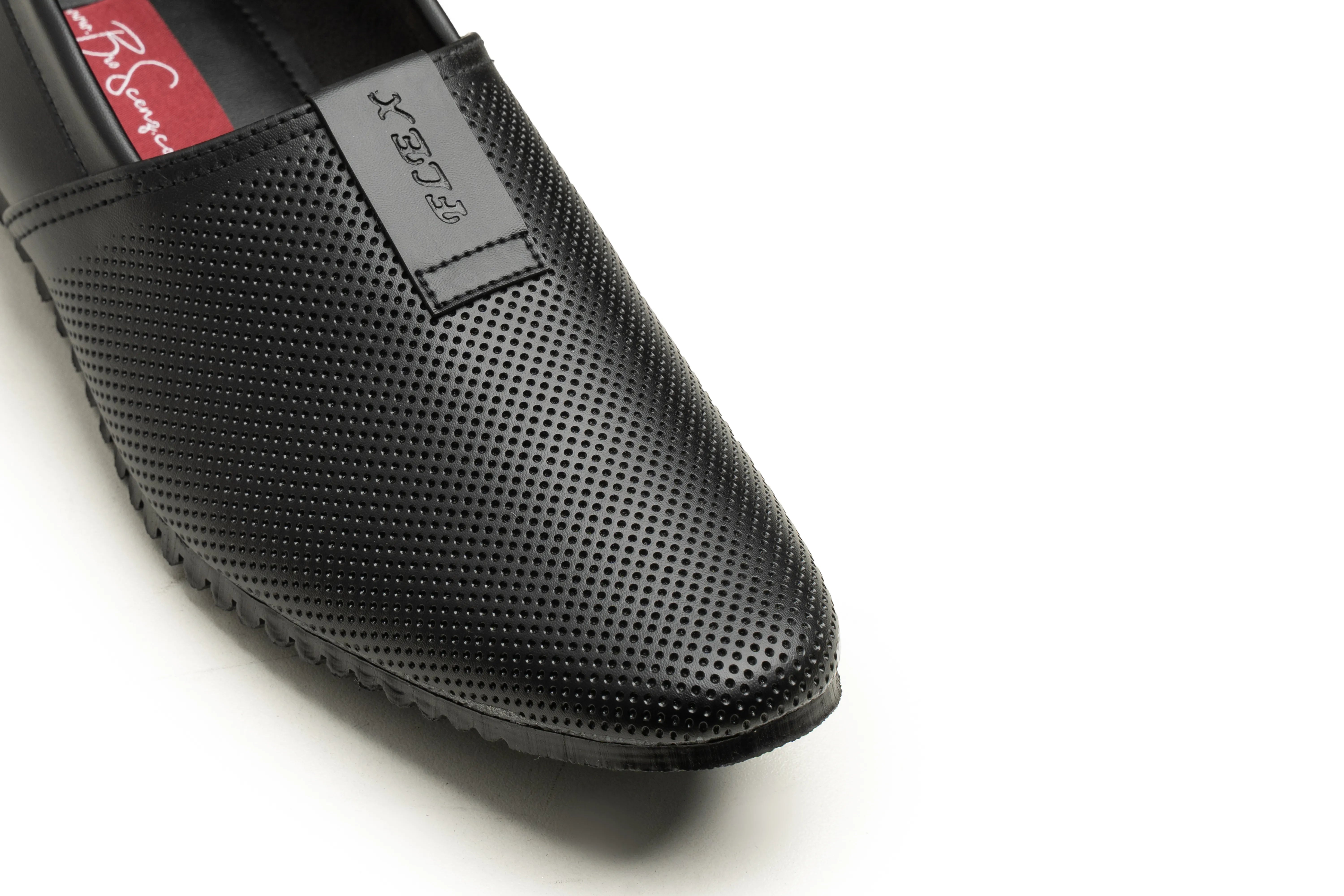 Close-up of a black slip-on shoe with perforated leather texture, a pointed toe, and a small gray brand tab on the side, photographed on a white background.
