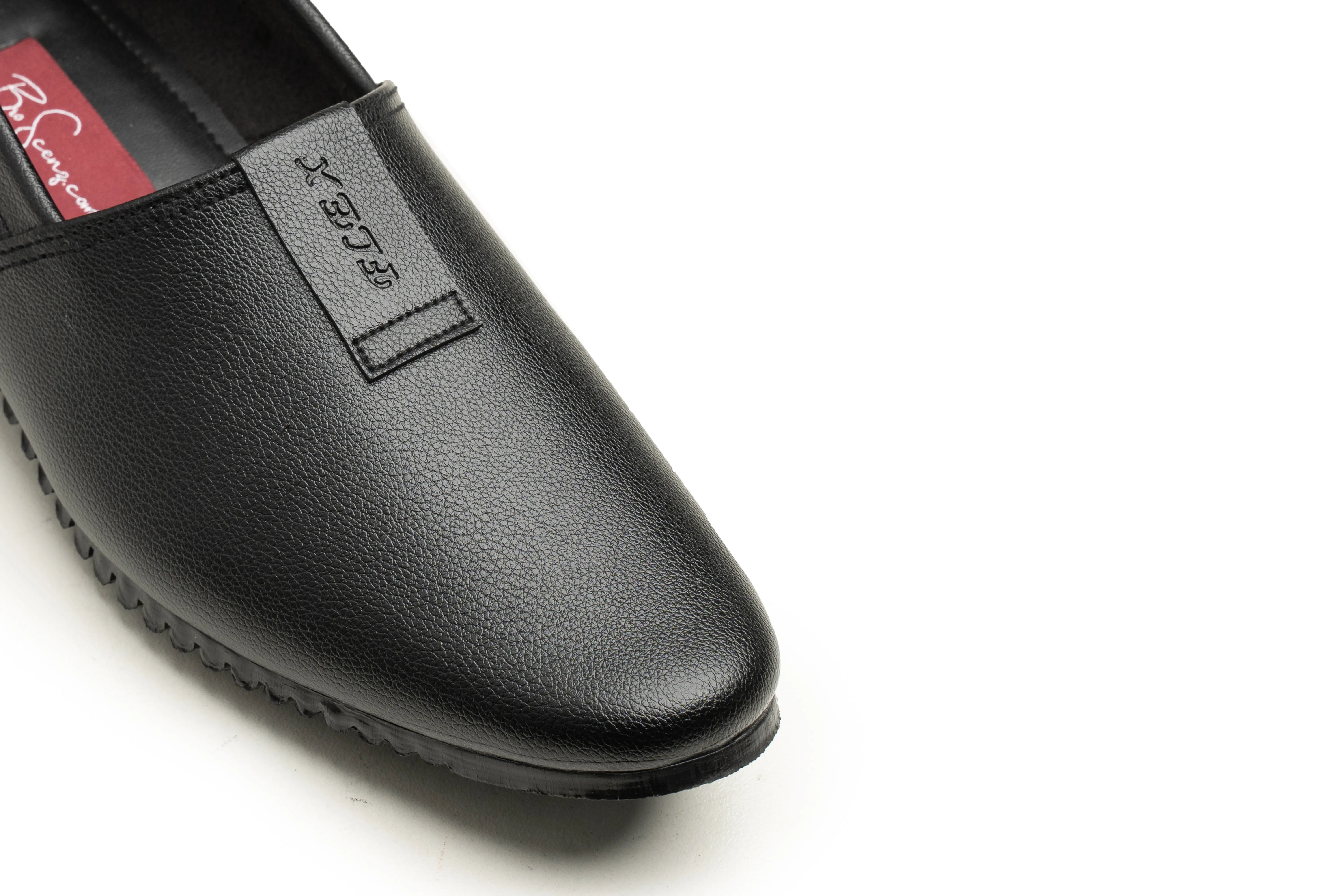 A close-up of a black leather slip-on loafer shoe, showing the textured toe, a small black brand tab on the side, and a red interior label with white text, set against a plain white background.