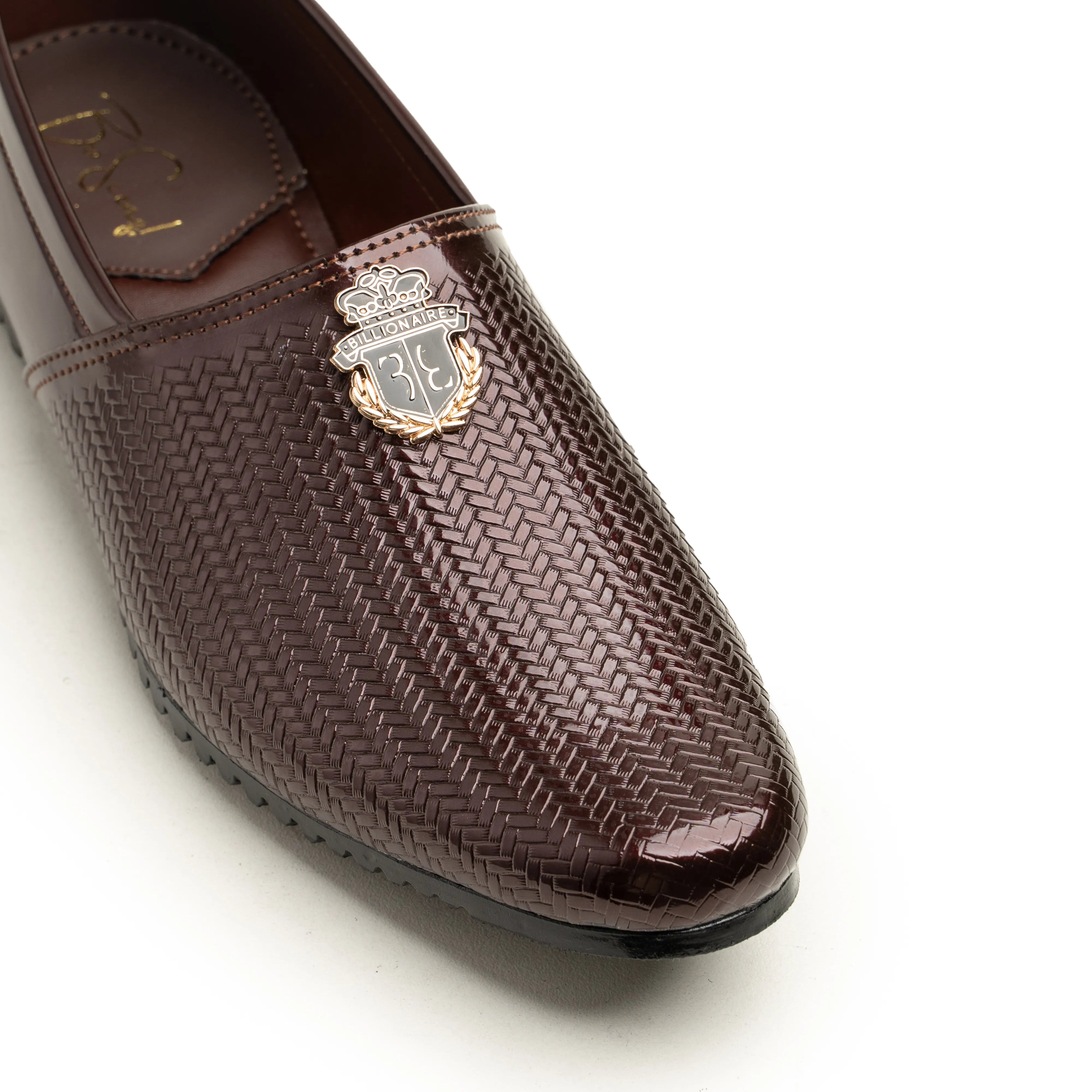 A close-up of a dark brown textured slip-on loafer with a metallic crest badge on the toe, set against a plain white background.