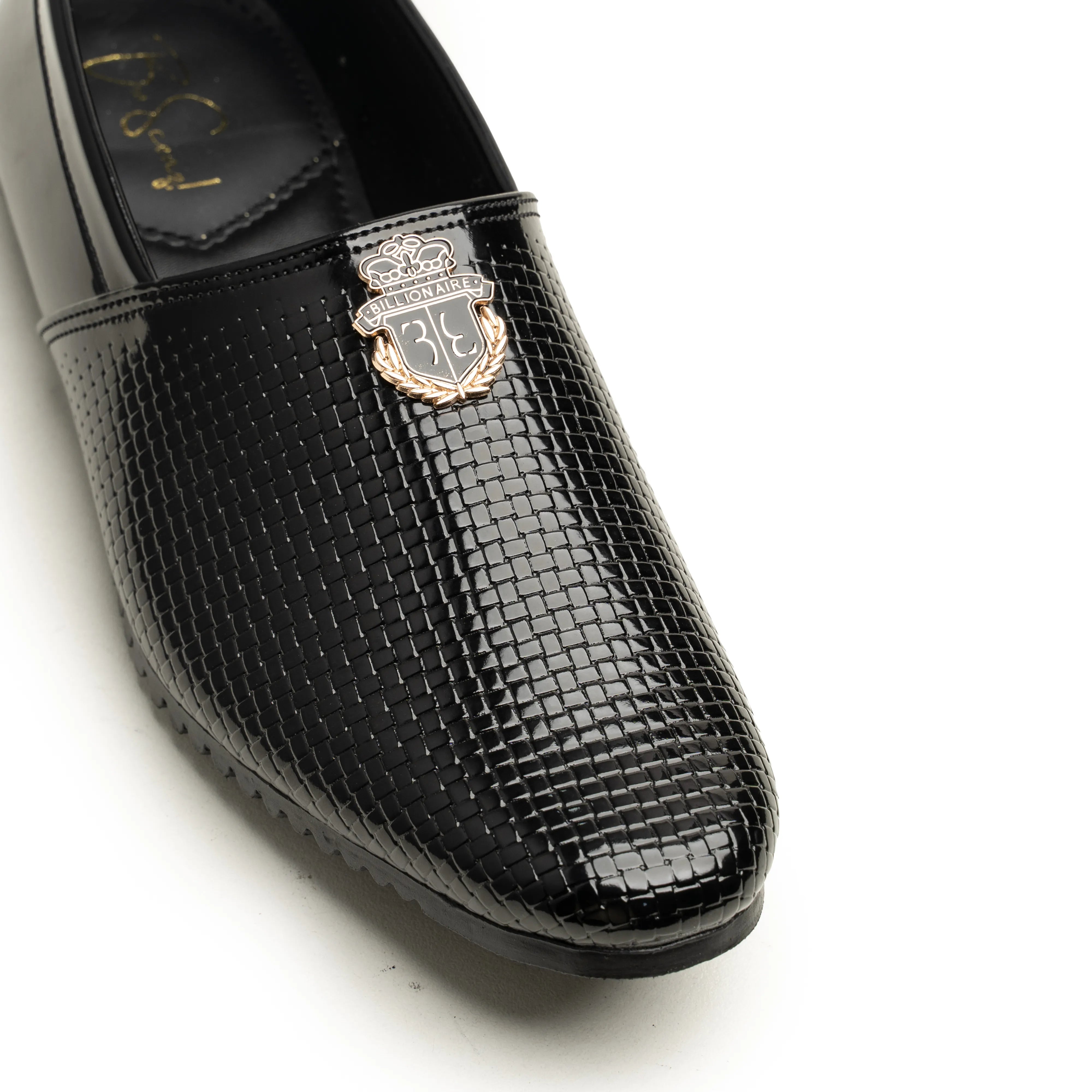 A close-up of a black slip-on loafer with a glossy crocodile-embossed texture and a decorative metallic crest badge on the vamp, photographed on a white background.